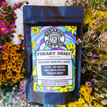A bag of Bizarre Coffee Freaky Deaky medium roast coffee. Tasting notes of golden raisins, brown sugar, cocoa nibs. Flowers in the background.