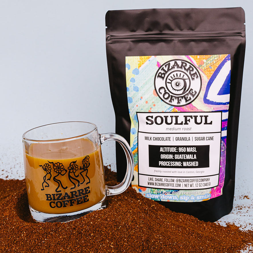 A glass mug that says Bizarre Coffee and displays dancing flowers in black while resting on top of coffee grounds and featuring a bag of Bizarre Coffee Soulful Medium Roast.