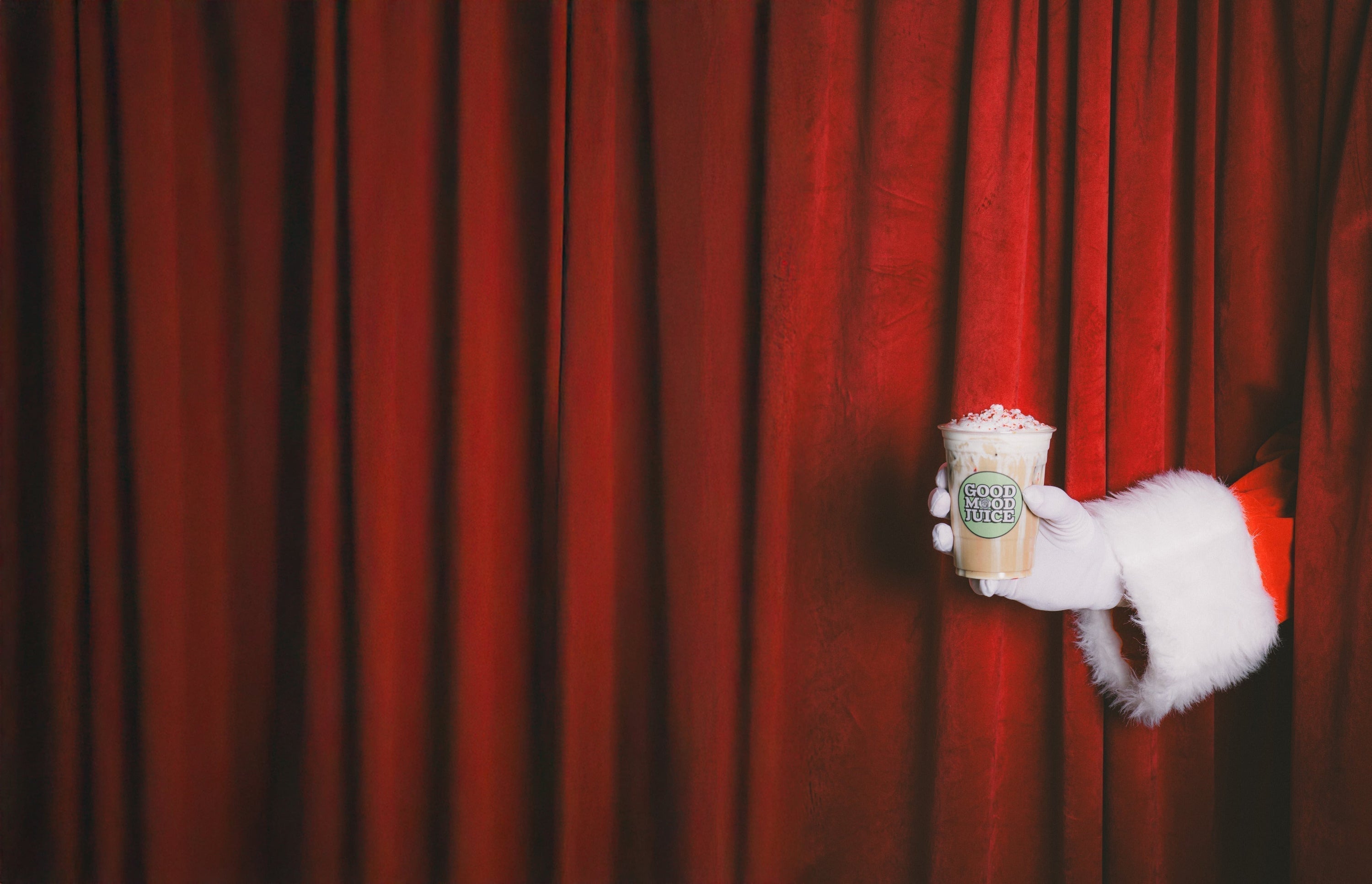 A person wearing a santa costume holding an arm in front of a red curtain with a drink in hand. 