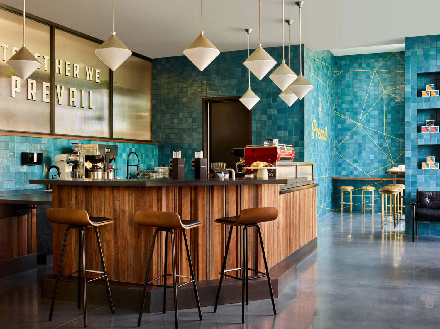 Modern interior of Prevail Coffee in Midtown Atlanta featuring a wood-paneled coffee bar, teal tile walls, pendant lighting, and the phrase “Together We Prevail.”