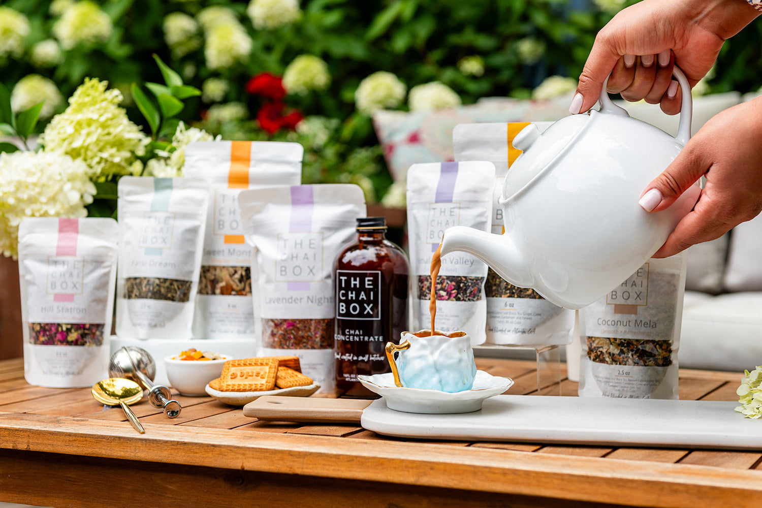 A close-up view of a wooden spoon scooping loose leaf chai tea blended with pink rose petals and green cardamom pods, sitting inside a glass jar.