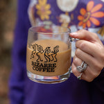 A glass mug  resting in a hand that says Bizarre Coffee and displays dancing flowers in black.