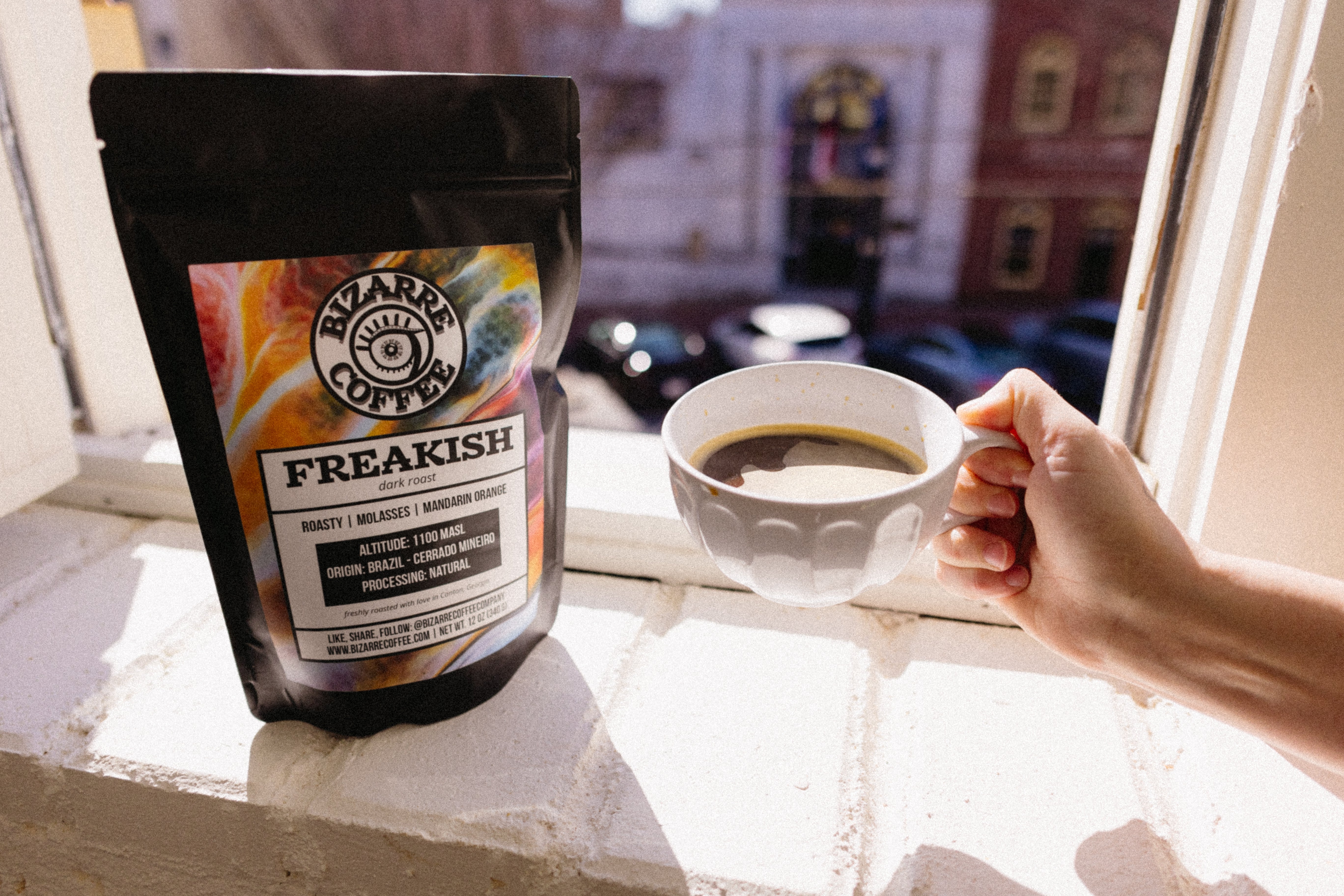 A bag of Freakish Bizarre Coffee with a mug in a person's hand.