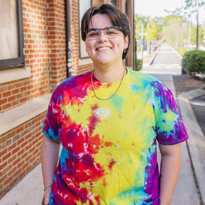 Person wearing a colorful tie-dye shirt standing on a sidewalk with a brick building in the background.
