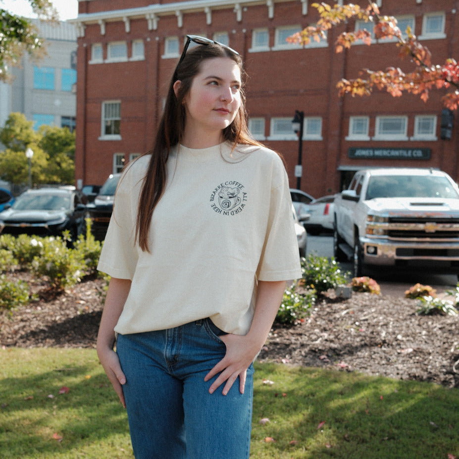 Person wearing a beige shirt and blue jeans standing outdoors with a building and cars in the background