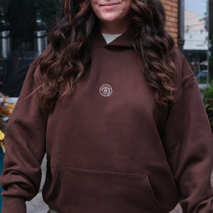 Person wearing a brown hoodie with a logo, standing outdoors.
