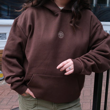 Person wearing a brown hoodie with a logo, standing on a sidewalk.